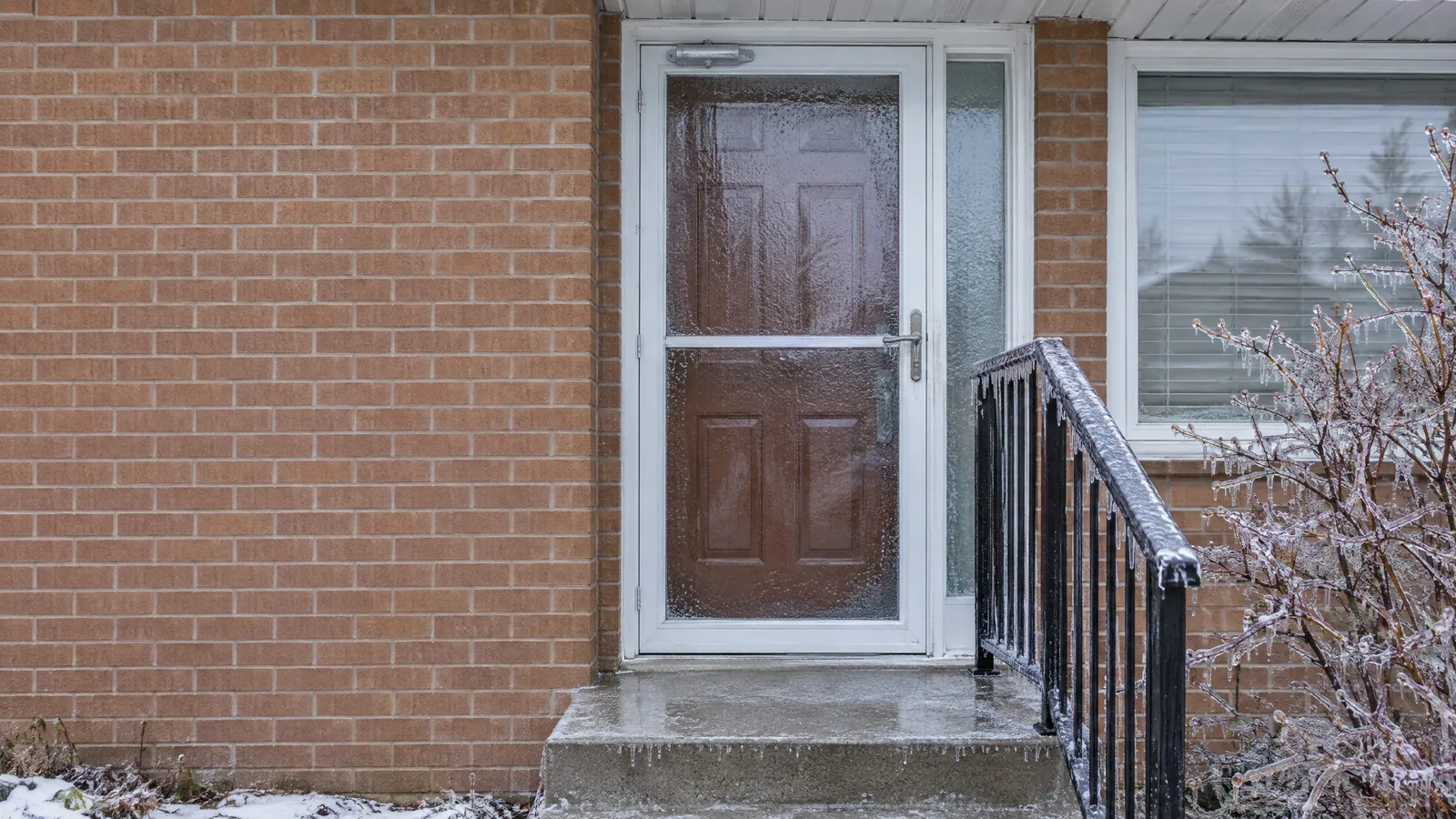 White full-view storm door glazed with ice from freezing rain in front of a brown panelled entry door